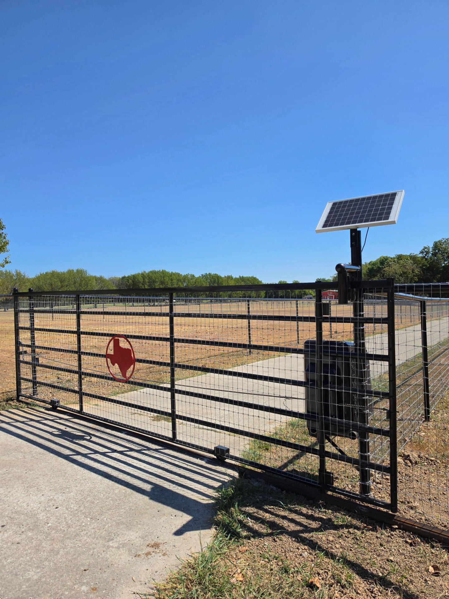 Black pipe rail ranch gate with a red Texas emblem and a solar-powered gate operator, opening onto a concrete driveway through a dry pasture.