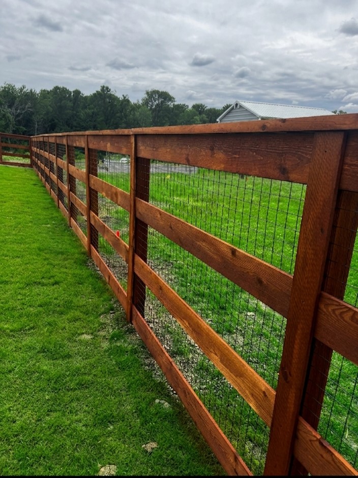 Stained cedar post-and-rail fence with four rails, a cap rail and black welded wire mesh on the inside, running along a green lawn with a gravel drive, outbuilding and tree line behind.