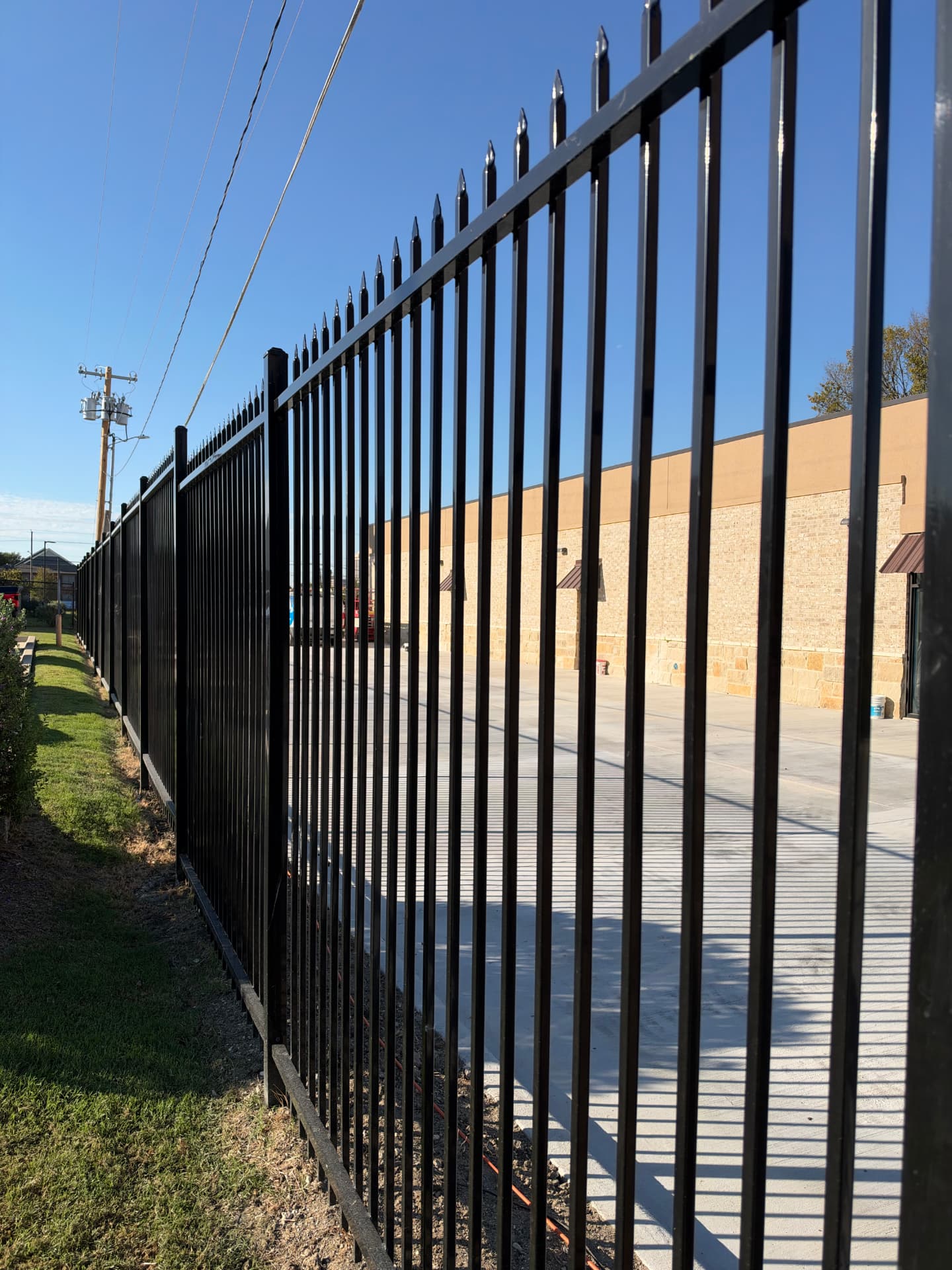 Tall black spear-top steel security fence separating a grassy boundary strip from a commercial building's concrete lot.