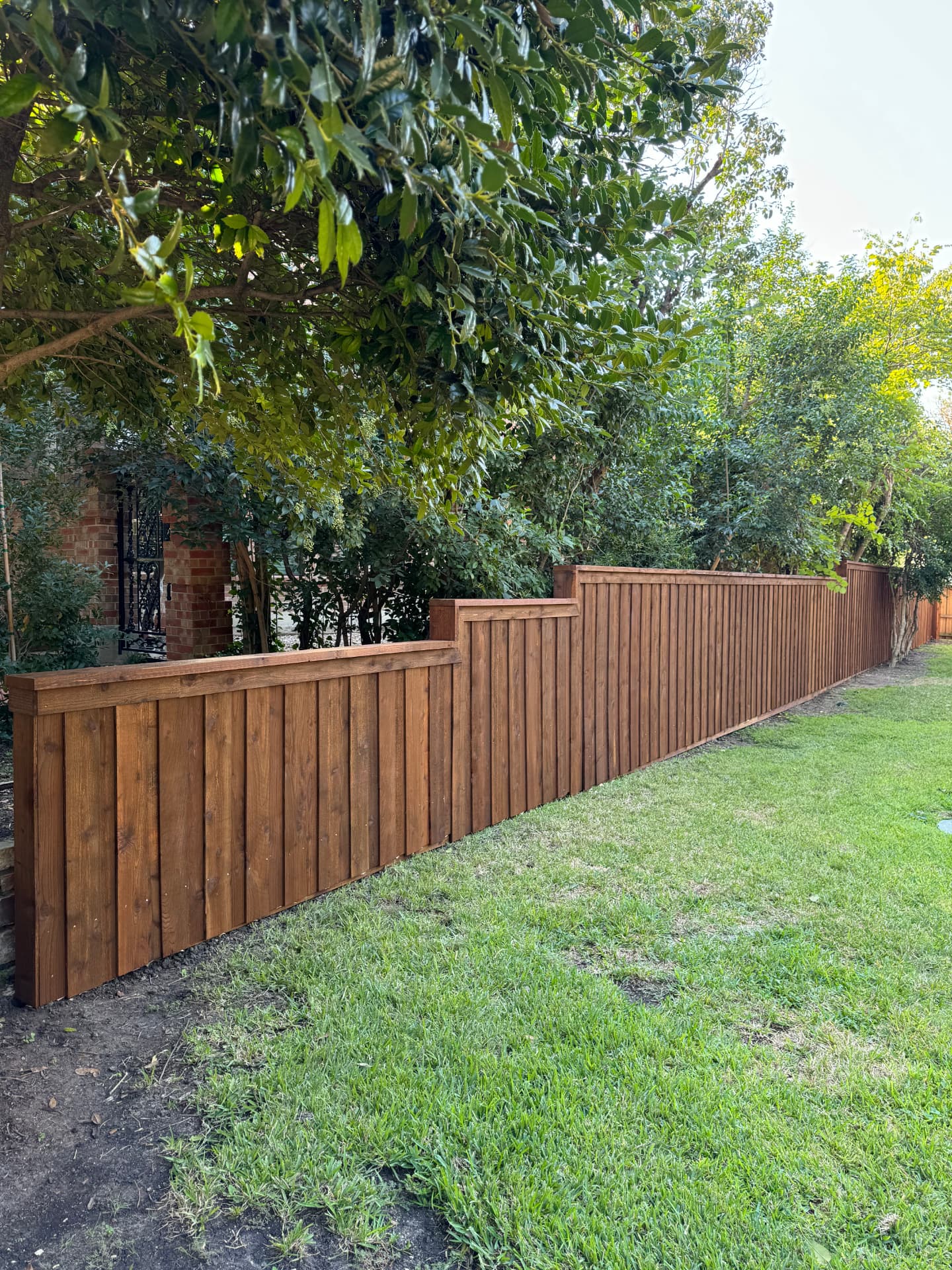 Vertical board cedar privacy fence with a cap rail and upper trim board, stepped upward in sections along a green lawn under mature trees.