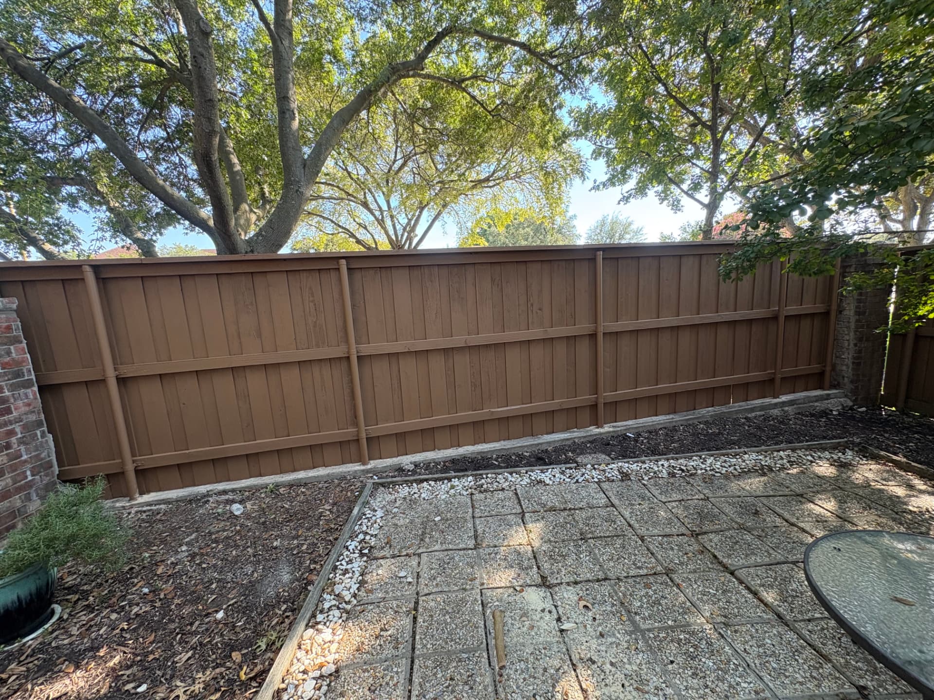 A freshly built, evenly stained brown wood privacy fence with a cap rail on steel posts, set between the brick pillars where the collapsed wall was, in front of a gravel patio.
