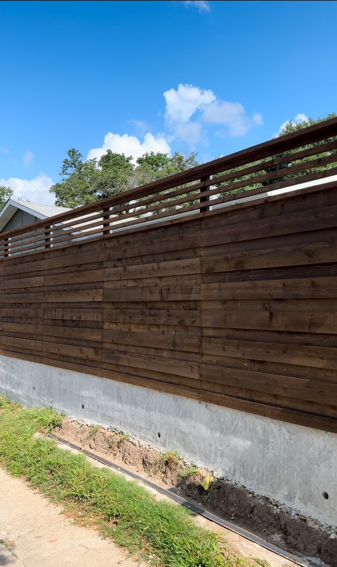 Tall horizontal cedar privacy fence in a dark chocolate stain with a narrower open-slat topper, mounted on a smooth poured concrete stem wall.