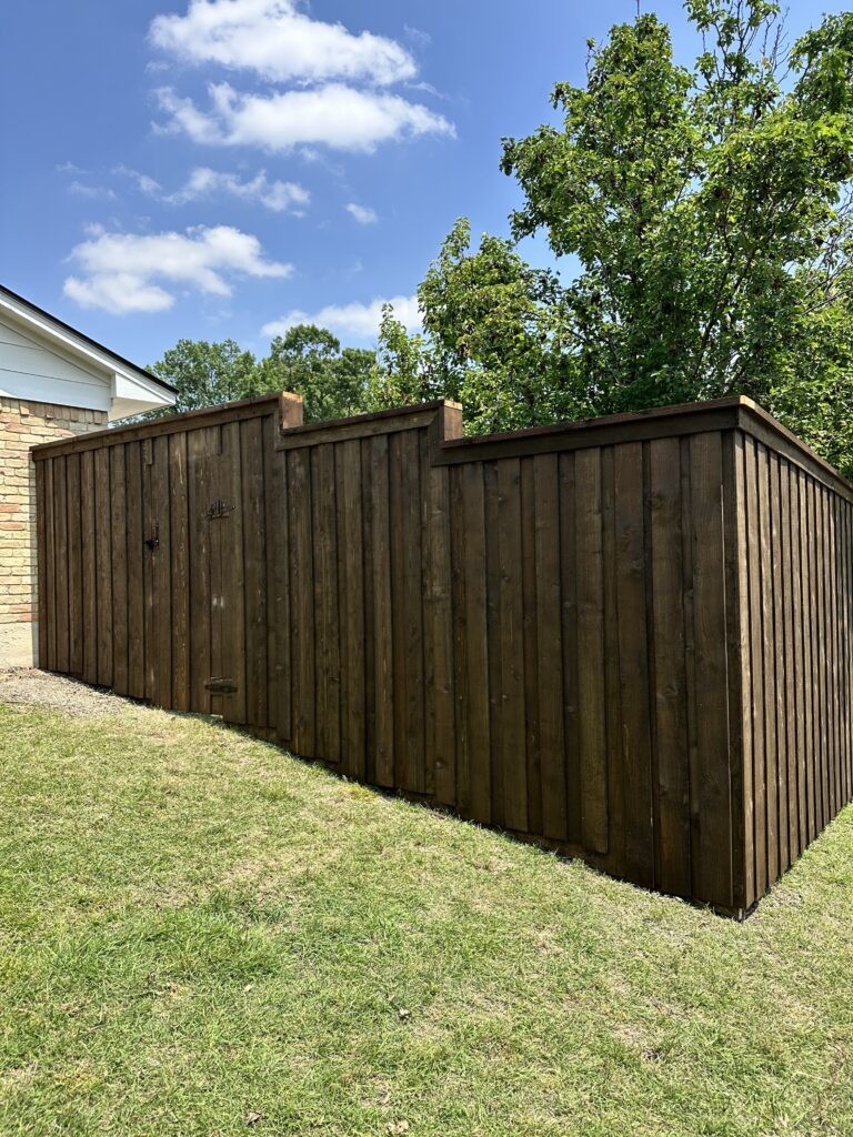 Stepped cedar privacy fence in a dark walnut stain with a cap rail and upper trim board, turning a corner on a sloped green lawn beside a light brick home, with a wood gate on black hardware.