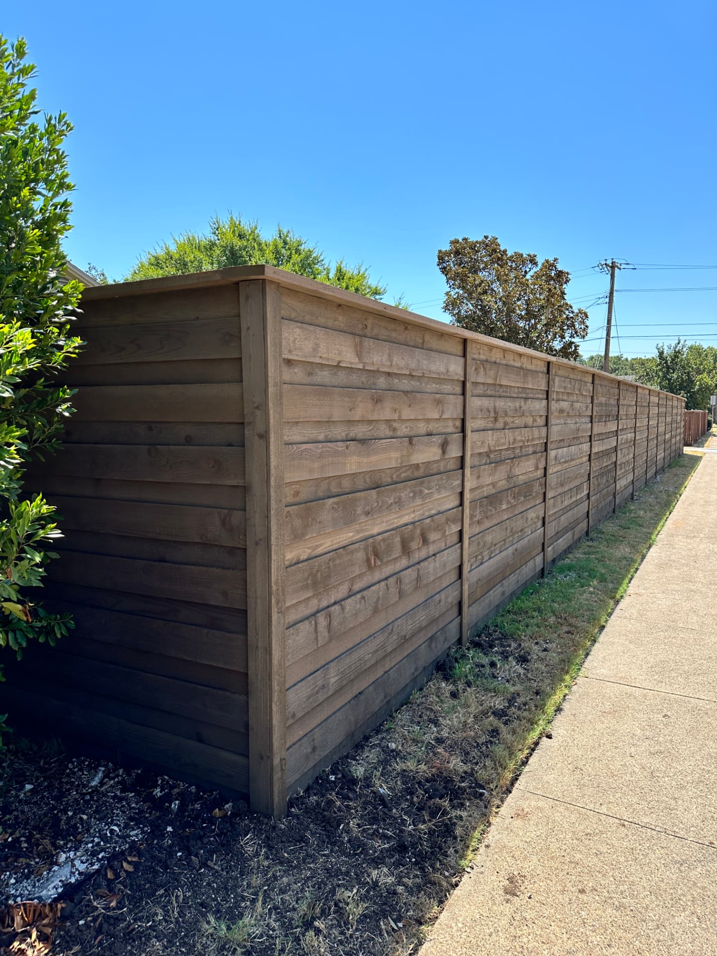 Horizontal cedar privacy fence in a muted walnut brown stain with vertical trim pieces and a flat top cap rail, running along a concrete sidewalk in front of a suburban home.