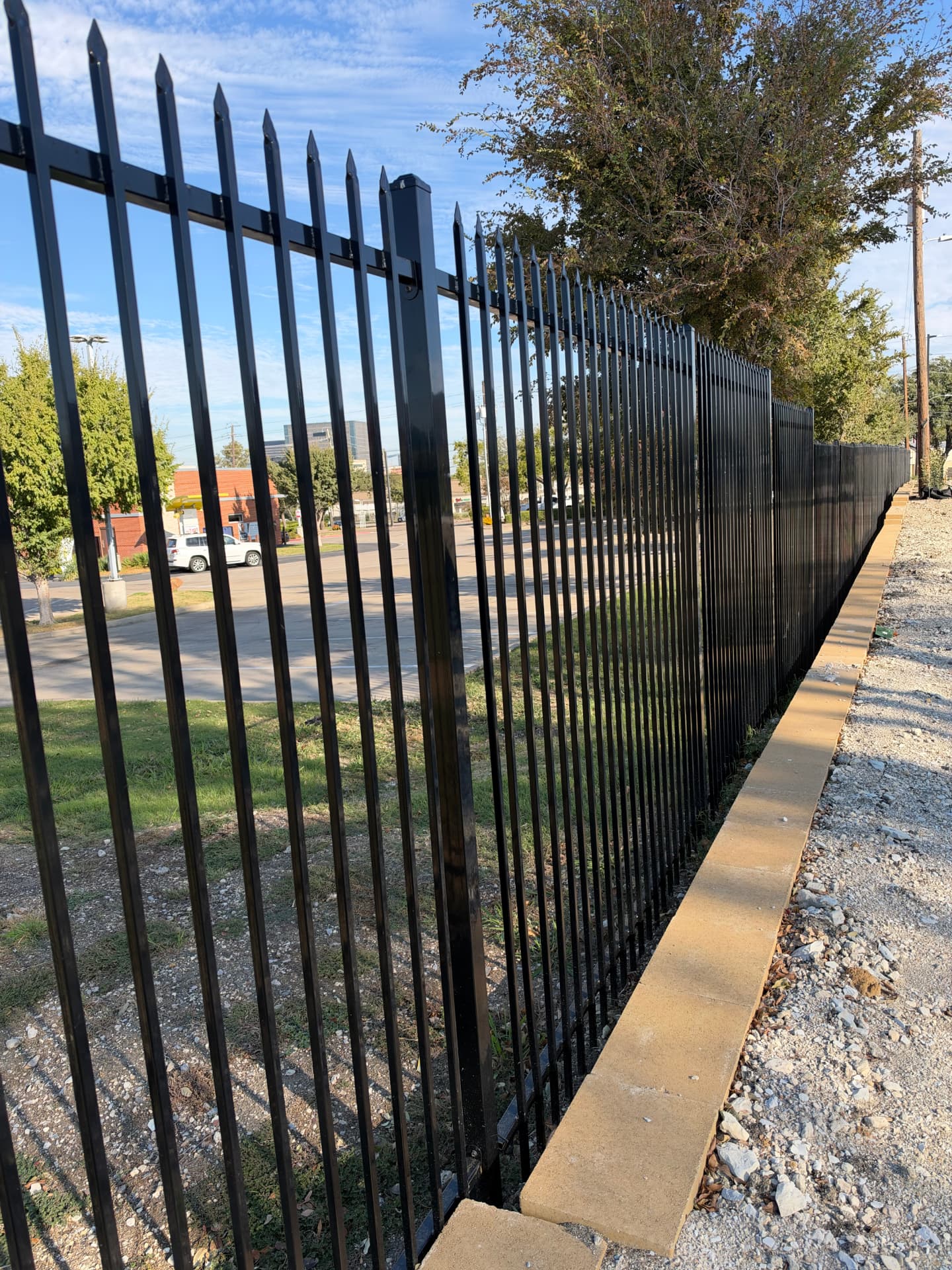 Black spear-top steel security fence with square posts running along the top of a stone retaining wall that borders a commercial parking lot.