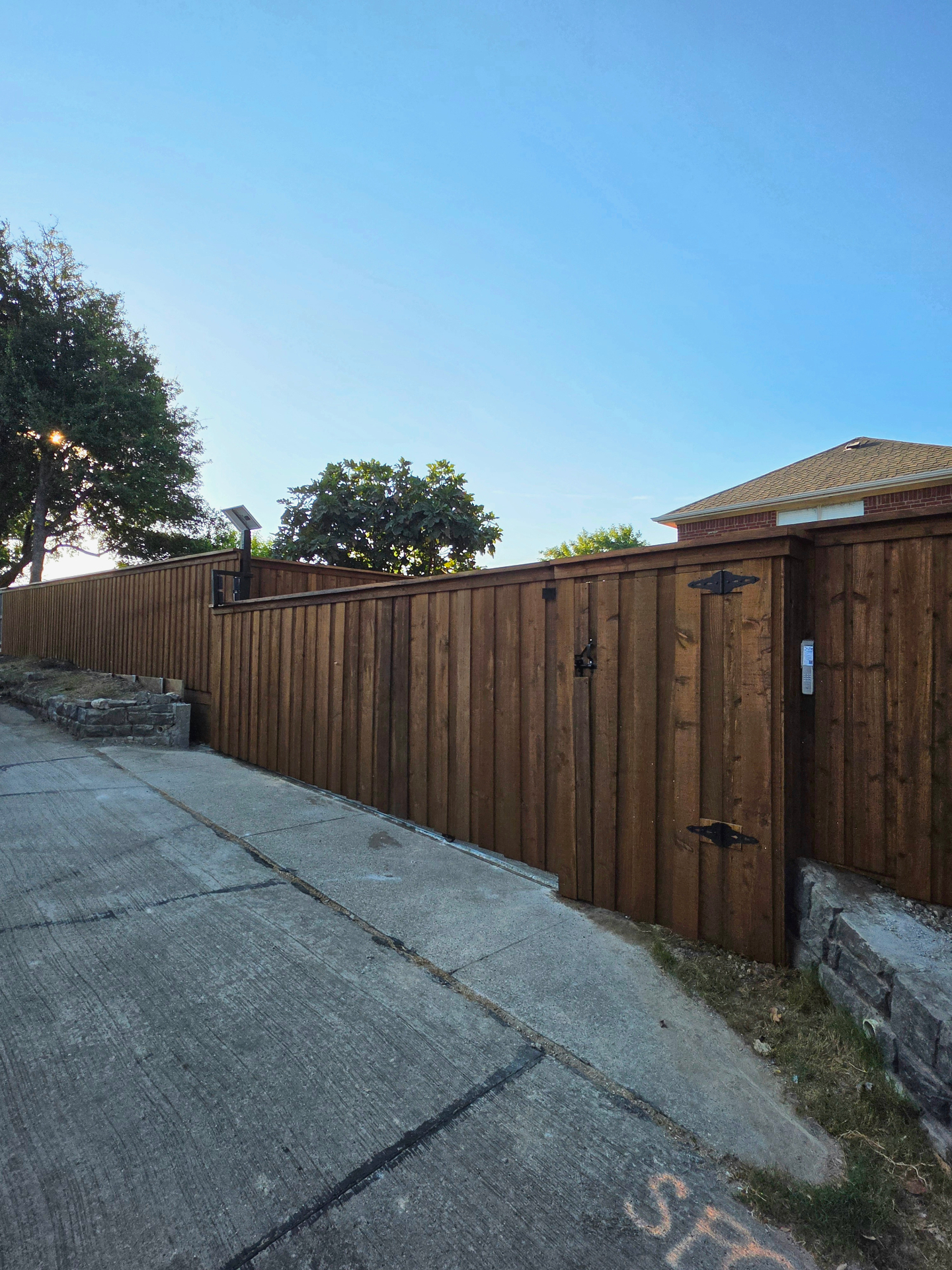 Board-on-board cedar sliding driveway gate with a cap, a matching pedestrian gate hung on black strap hinges with a keypad, and a small solar panel above the fence line, set in a scored concrete rear alley beside a stacked-stone wall.
