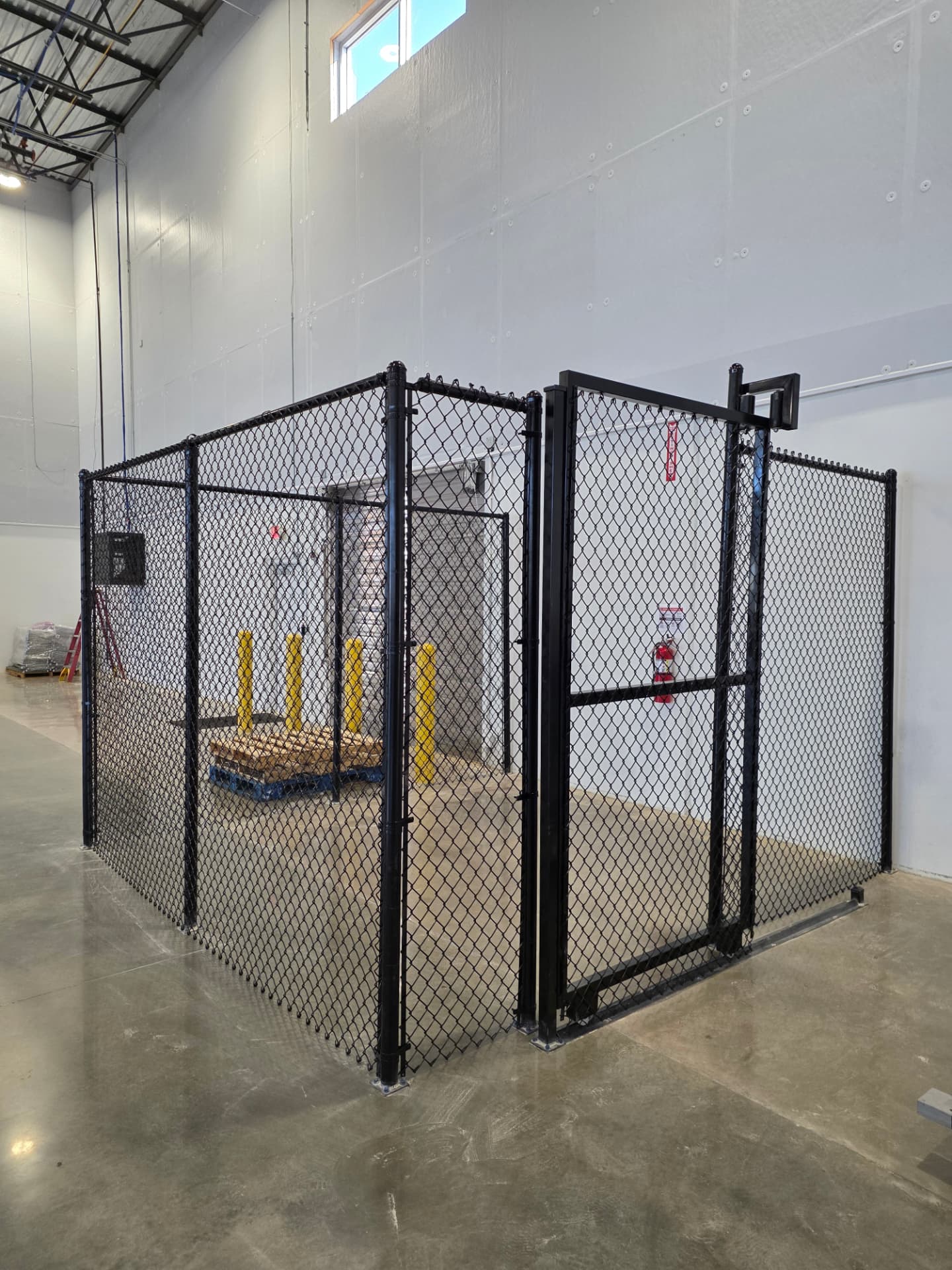 Black vinyl-coated chain link security cage with a full-height framed gate, built inside a warehouse on a polished concrete floor.