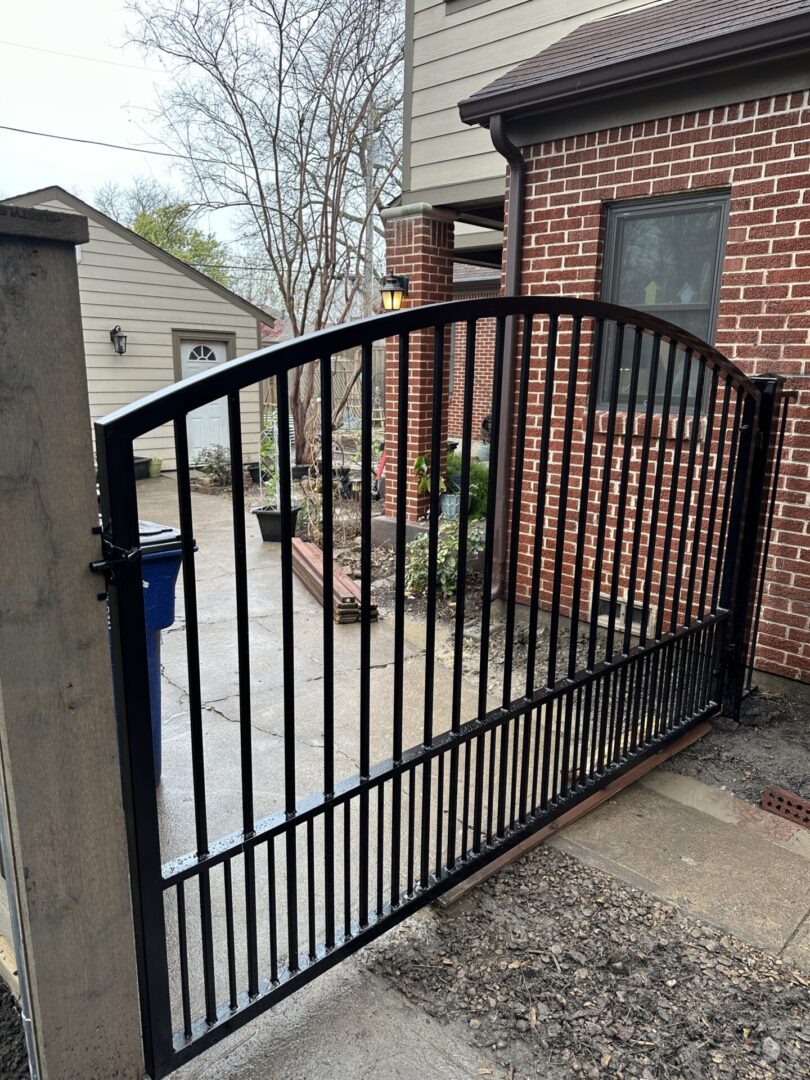 Arched black metal gate with closely spaced pickets along the bottom, hung between a wood post and a black steel post beside a brick wall.