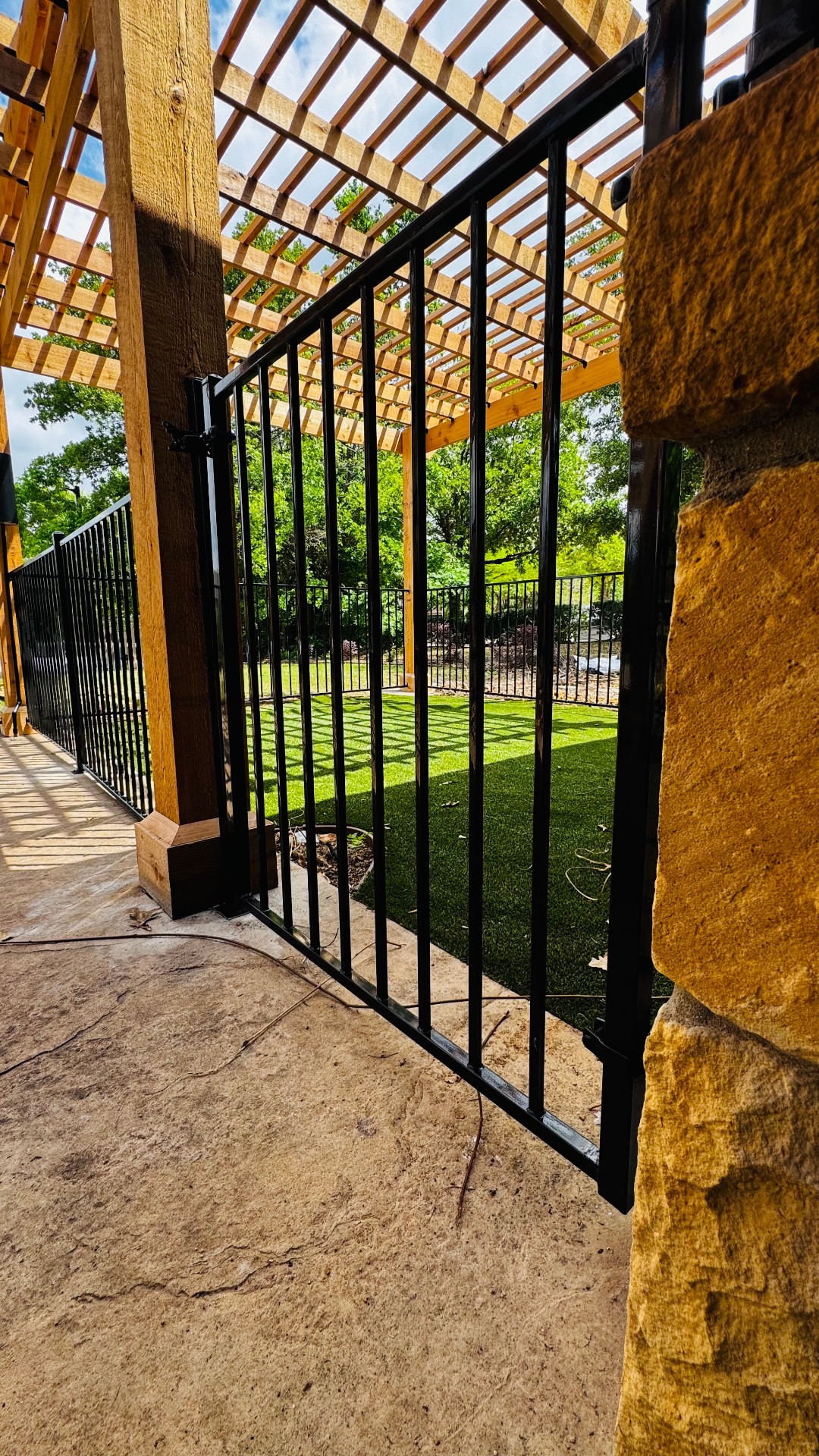 Glossy black metal swing gate with vertical pickets hung between a rough-faced stone pillar and a honey-brown pergola post, with matching black fence panels enclosing a green lawn behind.