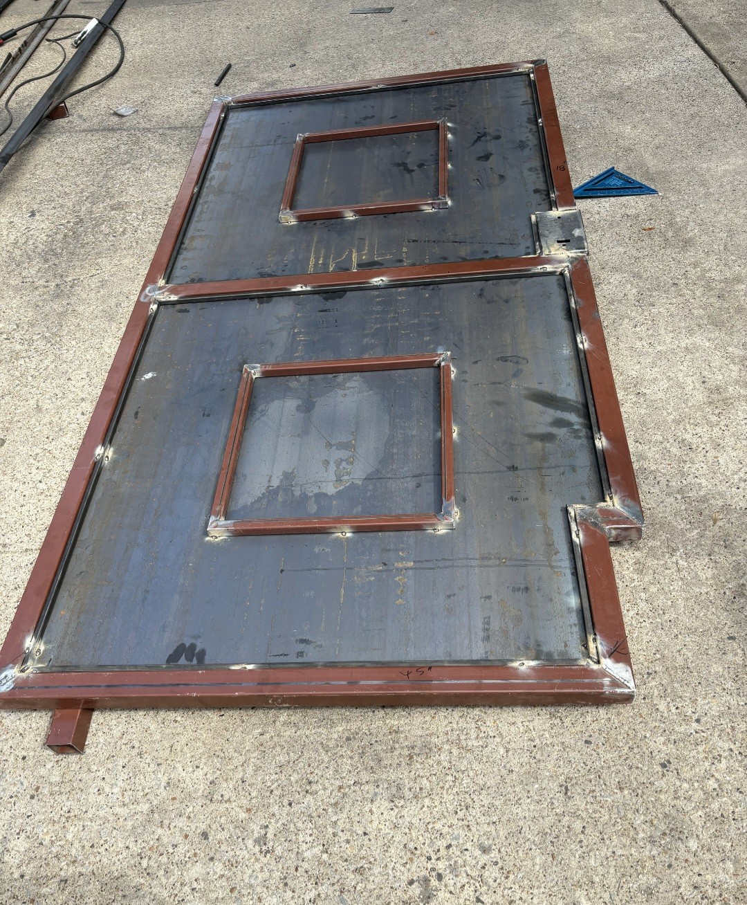 A custom steel gate panel under fabrication on the shop slab, showing square tubing framing, two inset trim boxes, a stepped cutout along the lower right edge, and tack welds over red primer.