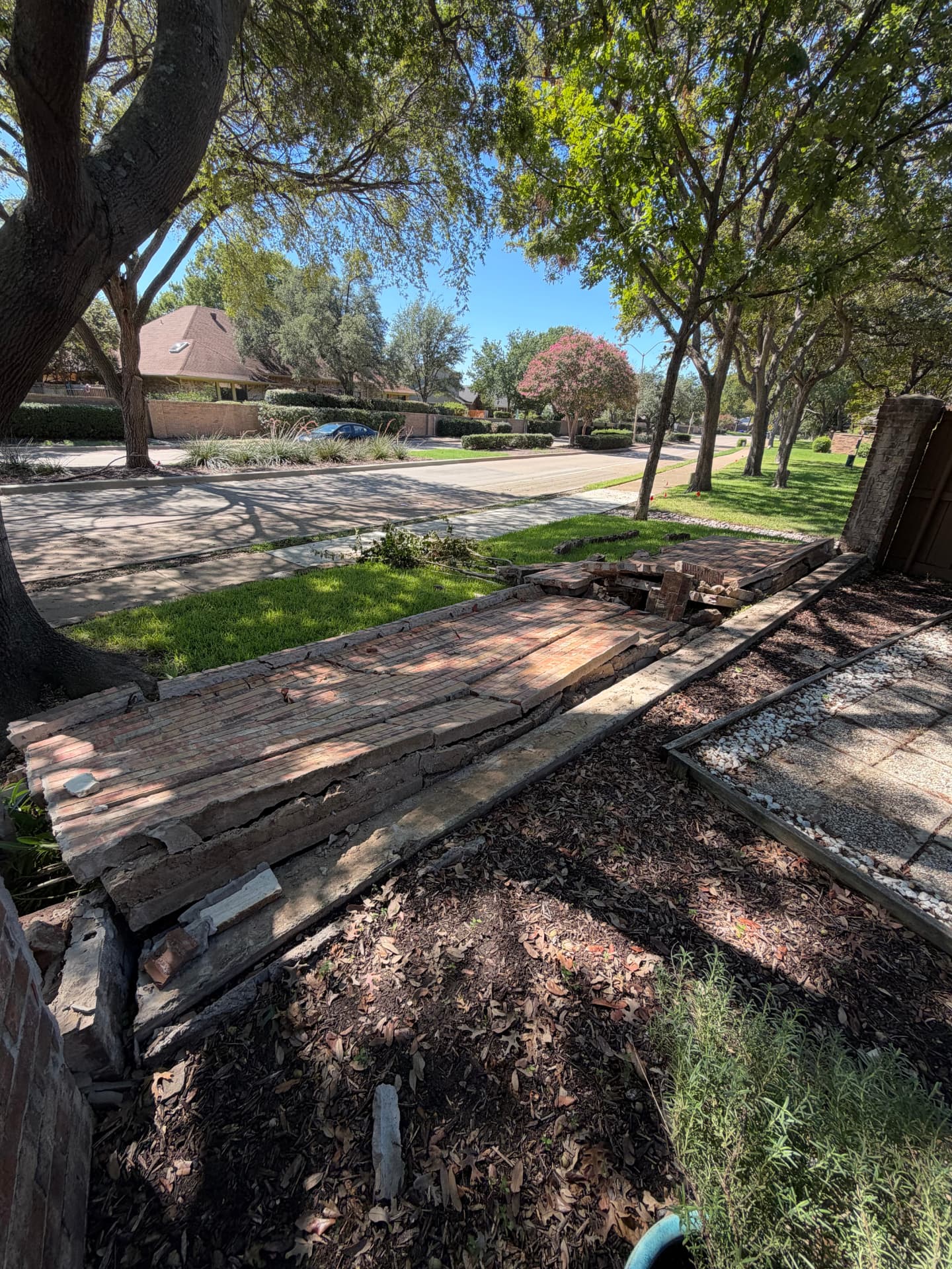 A brick perimeter wall that has collapsed entirely, lying in shattered slabs and loose bricks across the grass and curb line beside a mulched bed.