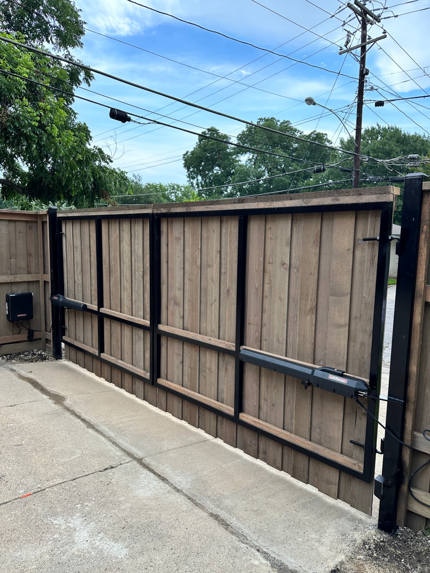 Double swing driveway gate clad in vertical cedar pickets on a black steel frame, fitted with two black LiftMaster swing gate operators, set in matching privacy fencing on a concrete drive.