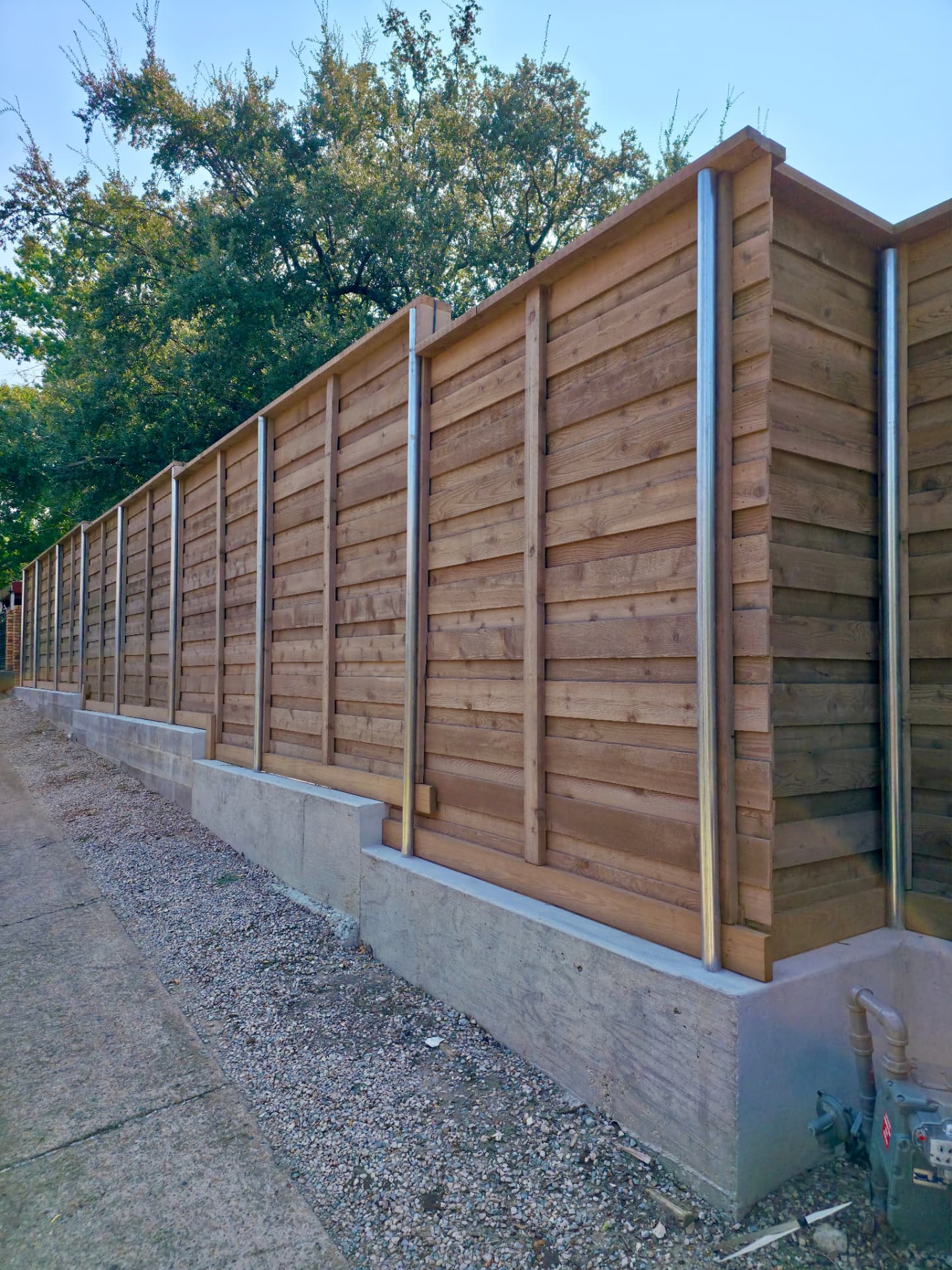 Horizontal cedar privacy fence in a warm golden-brown stain, built on round galvanized steel posts and stepped down along a poured concrete retaining wall, with trees behind.