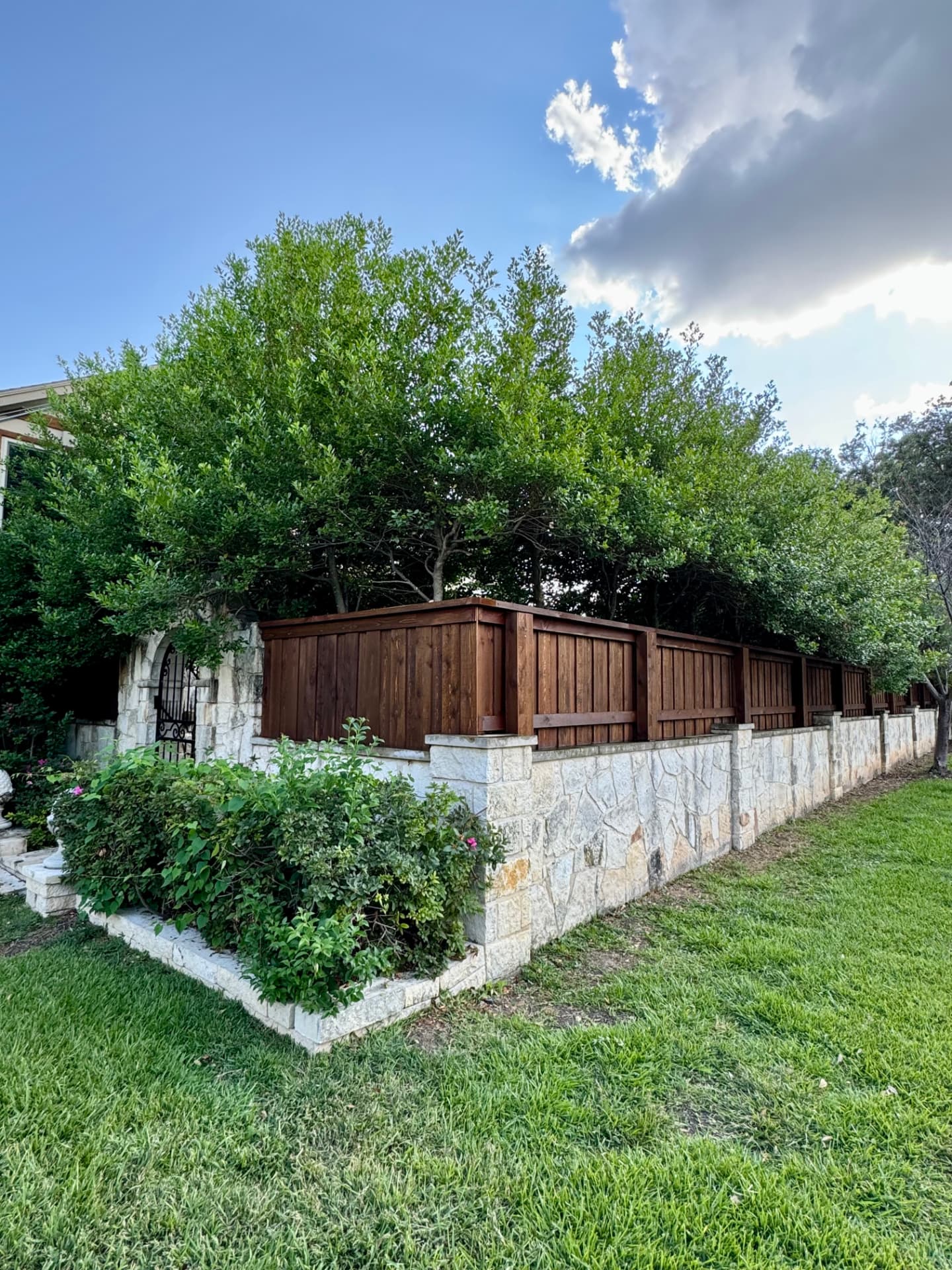 Squared natural stone wall and pillars topped with dark stained cedar privacy panels, with an arched stone entry fitted with a black iron gate.