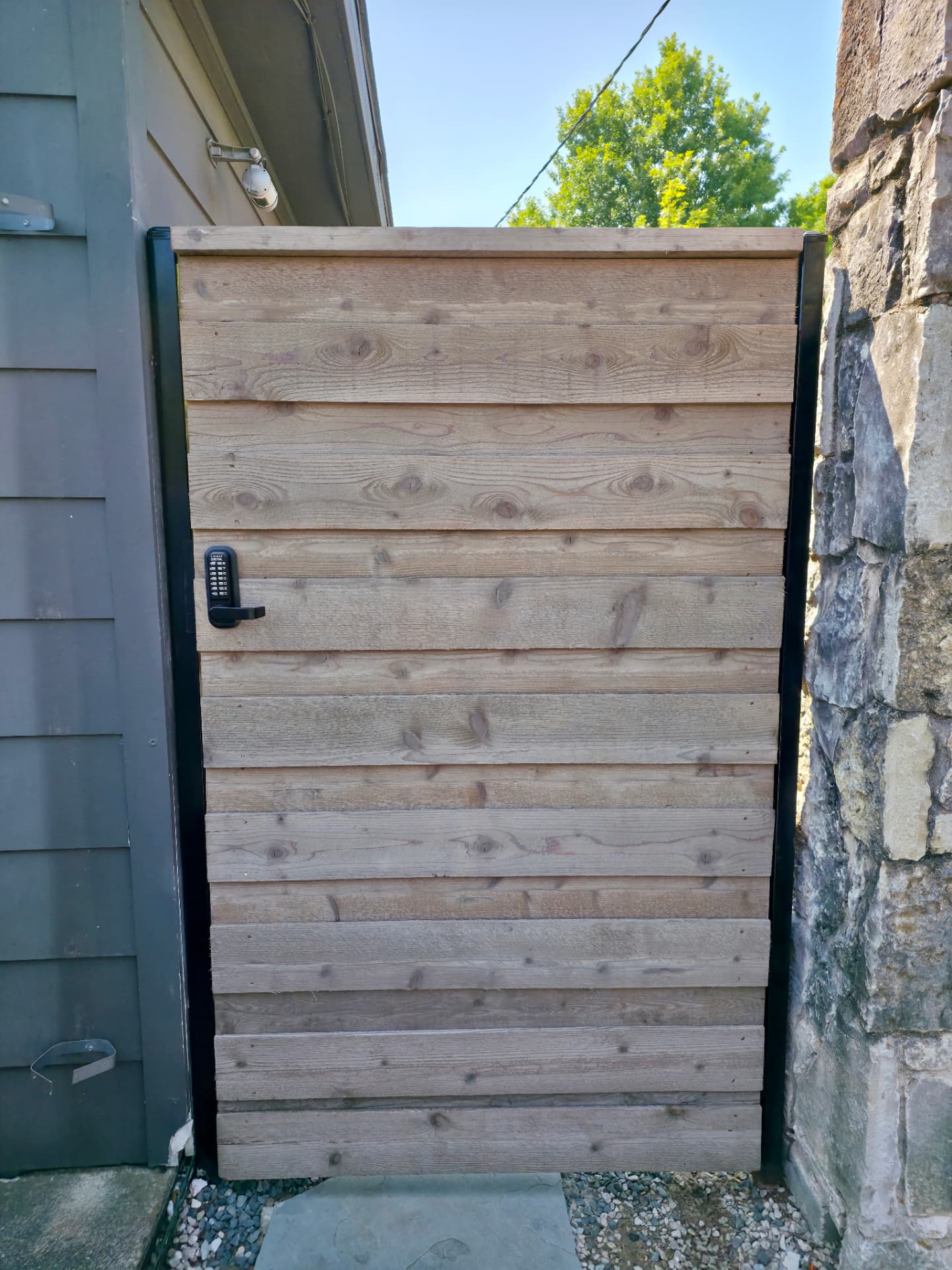 Close view of a horizontal cedar pedestrian gate in a weathered natural stain with a flat cap board and a black keypad lock, set between a dark-sided building and a rough stone pillar over flagstone pavers.