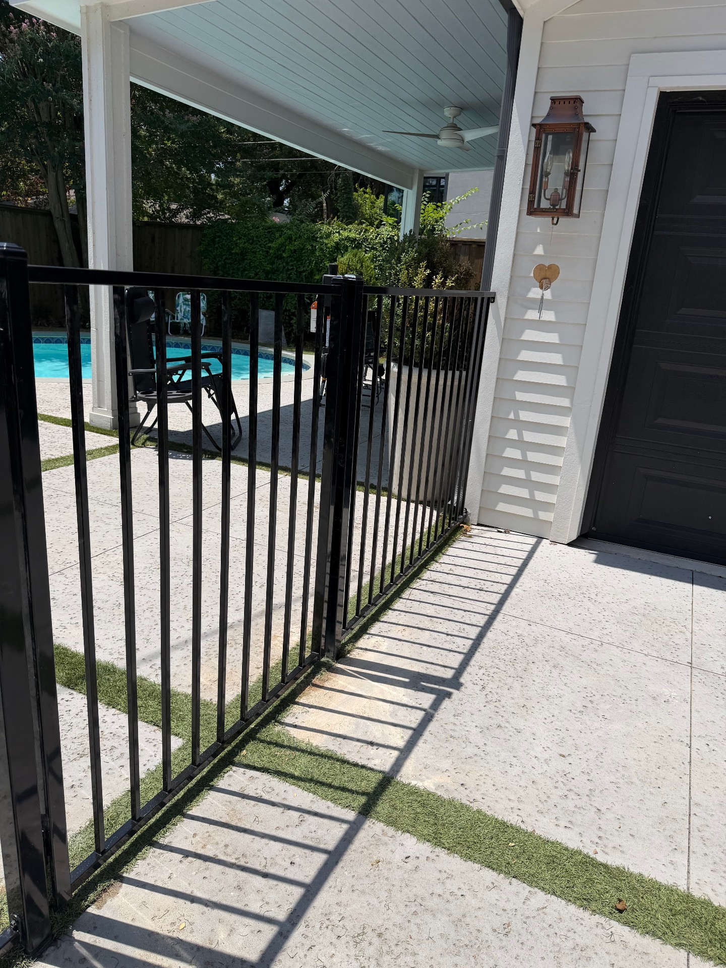 Gloss black flat-top ornamental fence with vertical pickets and an integrated gate surrounding a pool patio paved with concrete pavers and turf strips.
