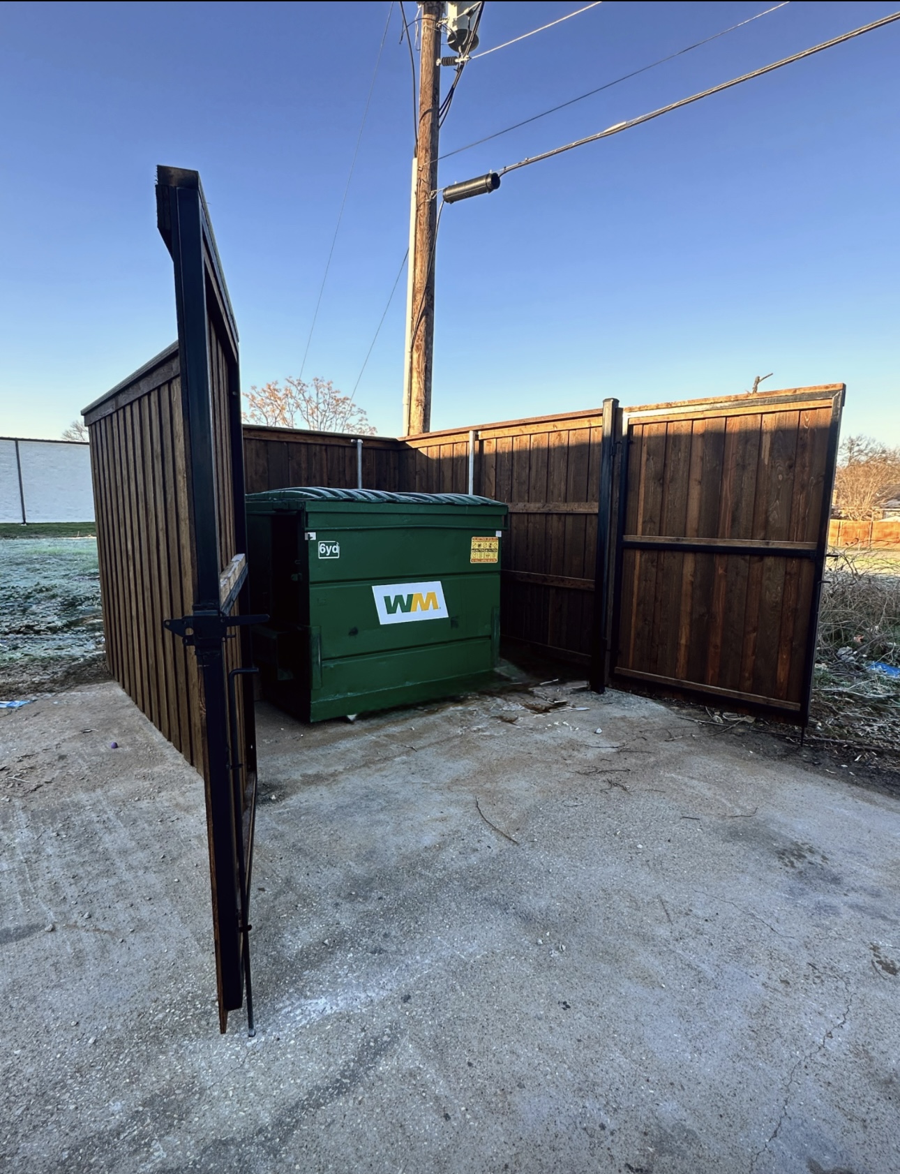 Stained wood dumpster enclosure with black steel frames and double swing gates standing open around a green roll-off container on a concrete pad.