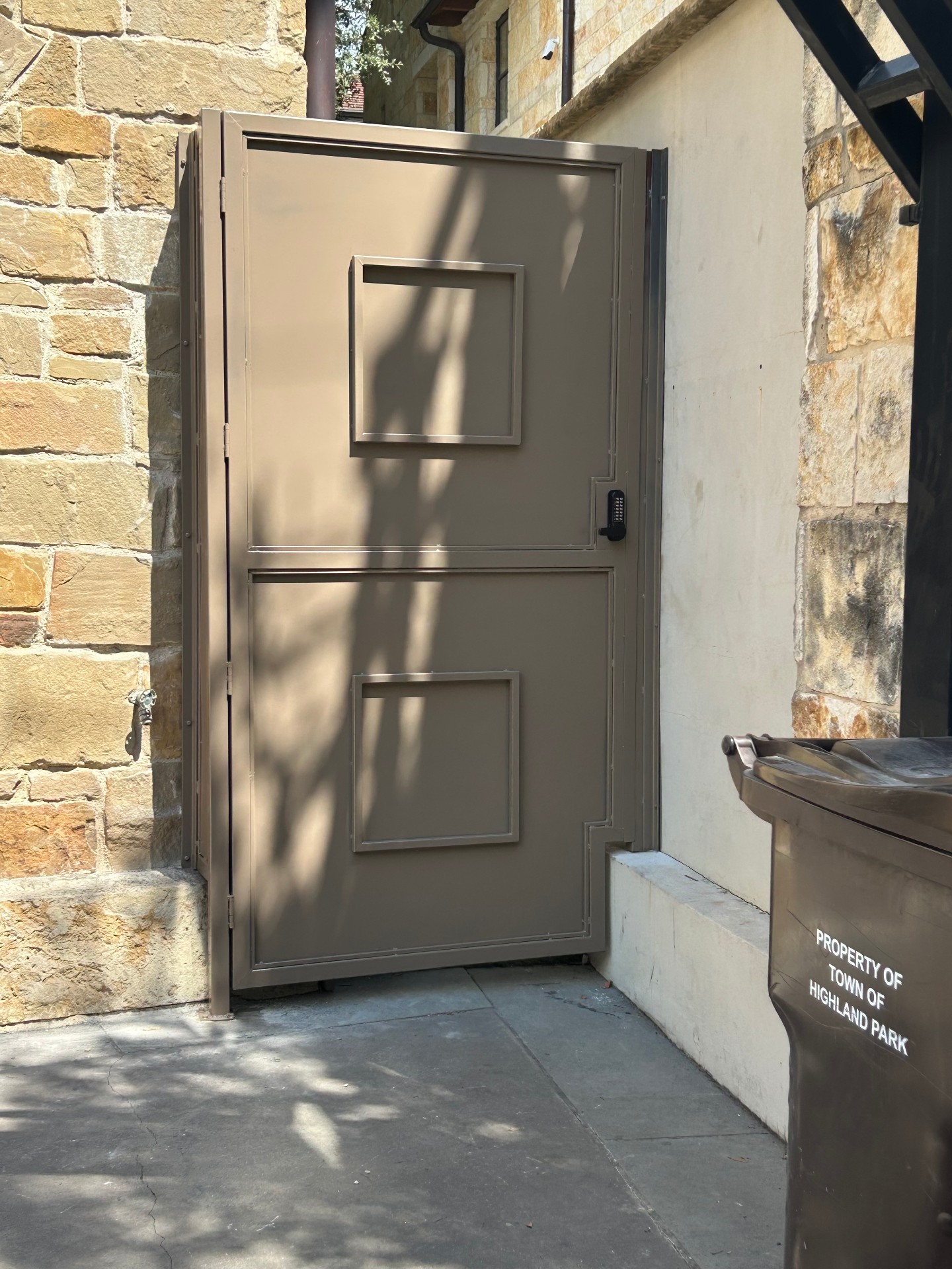 The finished custom gate in matte taupe with two raised inset panels and a stepped cutout over a concrete curb, hung on barrel hinges with a black keypad lock, between a stone wall and a stucco wall.