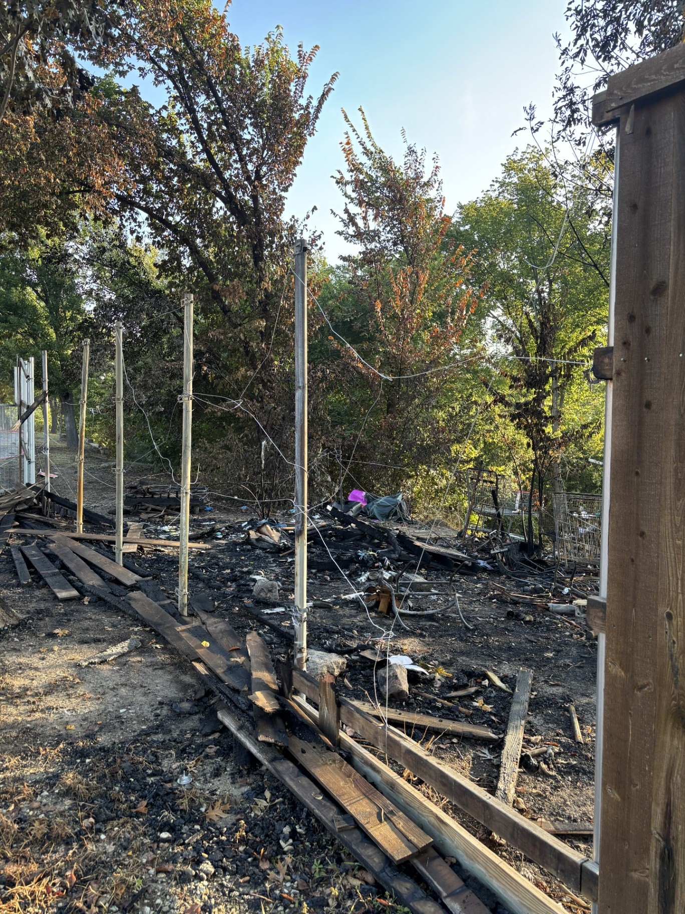 A fence line burned down to its round steel posts: pickets and rails gone from the frame, charred boards and black ash along the ground, scorched brush behind, and mounting brackets still on the upright posts.