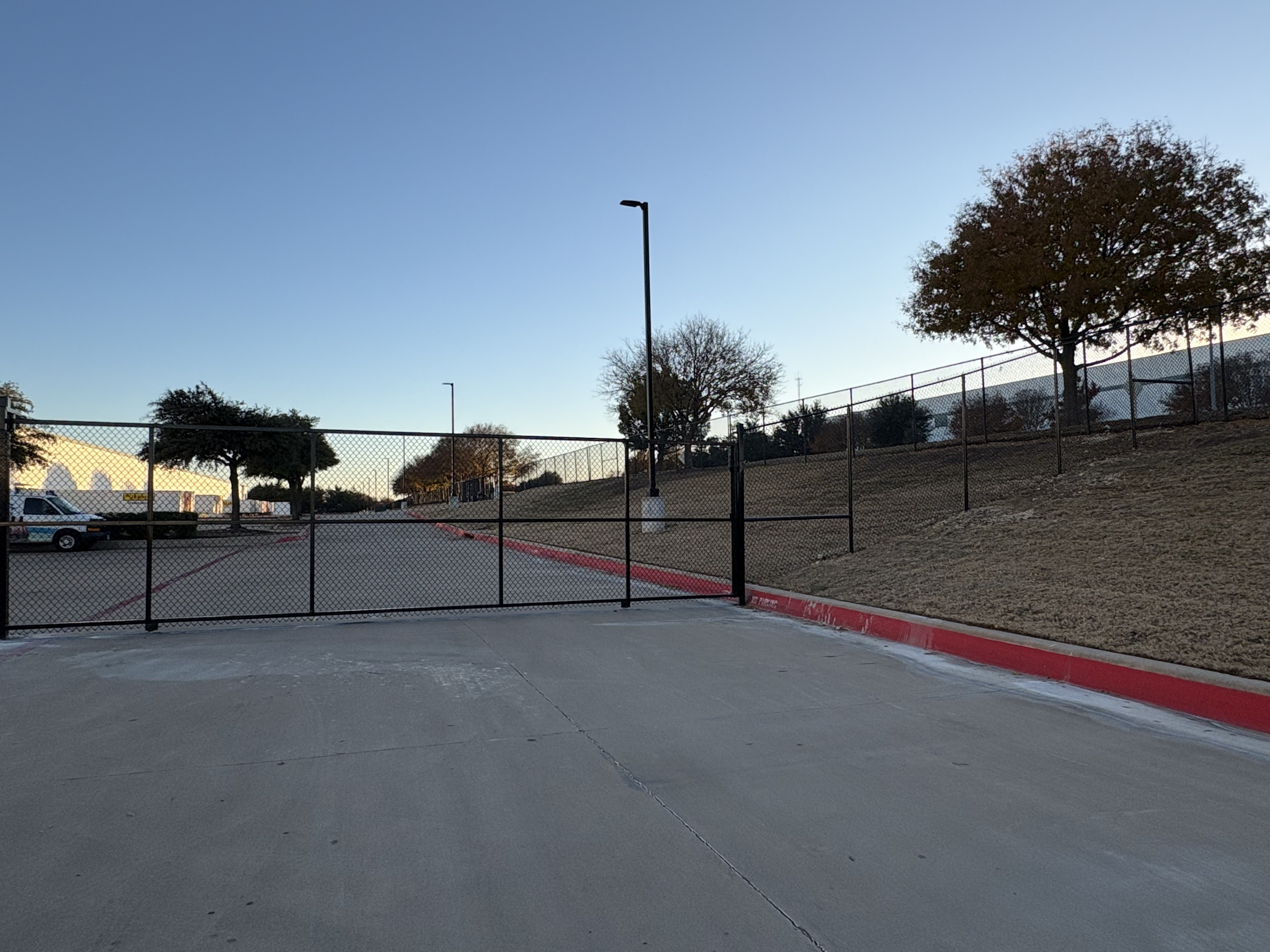 Black vinyl-coated chain link commercial fence with a wide black rolling drive gate spanning a concrete entrance road, red fire-lane curbs and a grassy berm behind, a warehouse building off to the left.