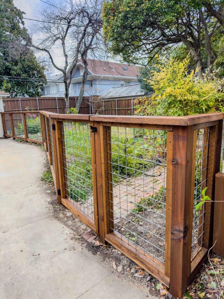 Stained wood-framed hog wire panels with a swinging gate hung on black hardware and a flat cap rail, beside a concrete driveway and a landscaped garden.
