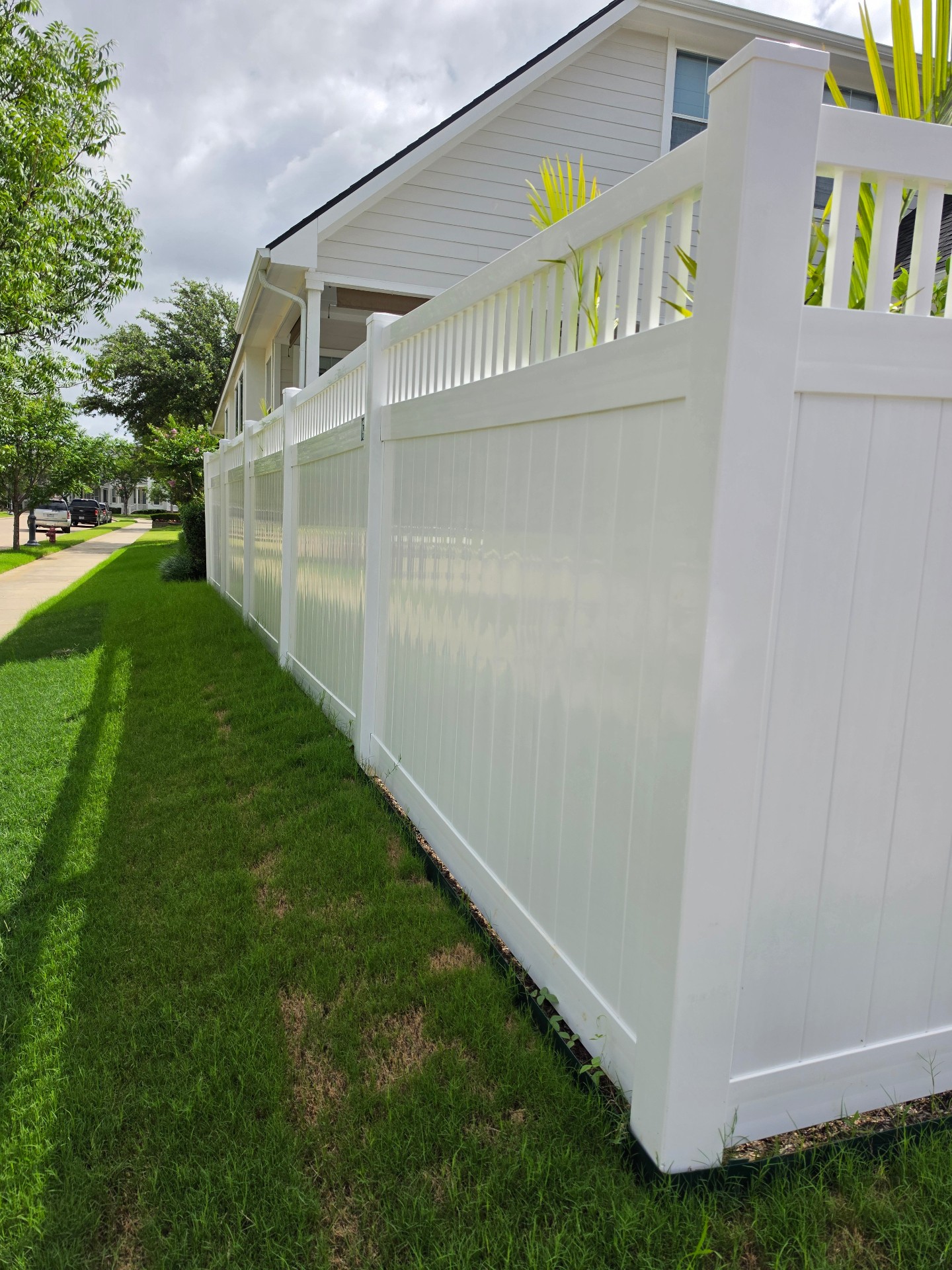 Bright white vinyl privacy fence with tongue-and-groove lower panels, capped posts and an open vertical spindle topper, running along a green lawn in front of a home with light horizontal siding.