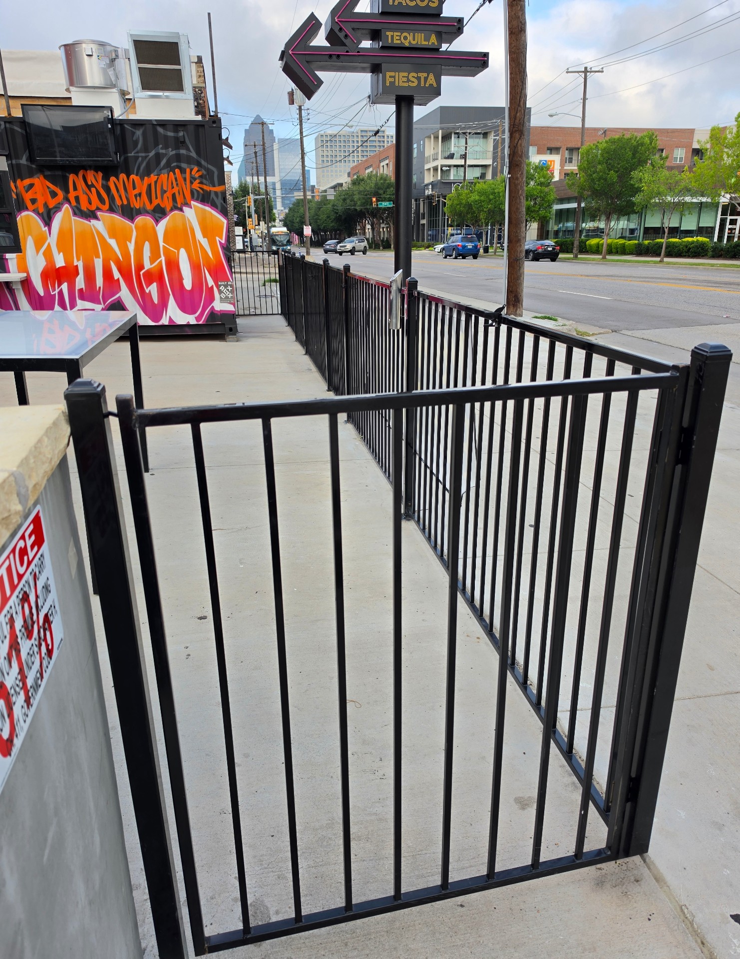 Open black metal pedestrian gate with barrel hinges and square post caps on a long run of matching vertical picket perimeter fencing along a concrete sidewalk patio outside a restaurant.