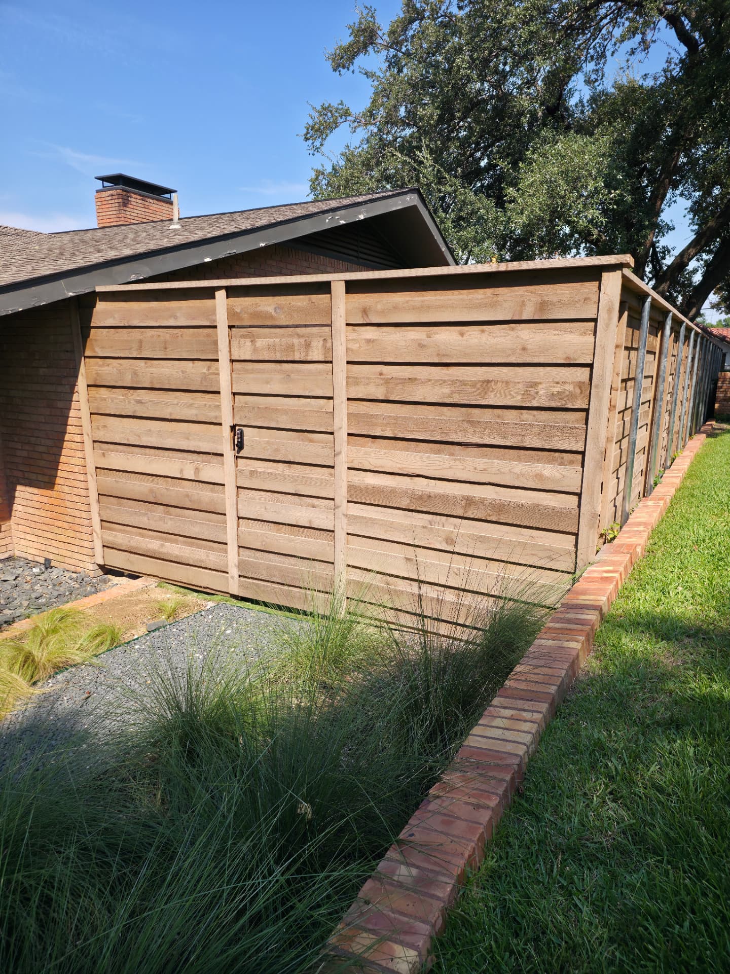 Horizontal cedar privacy fence in a golden-brown stain with a flat cap board and a matching gate fitted with a black keypad latch, beside a red brick home with a gravel bed of ornamental grasses.