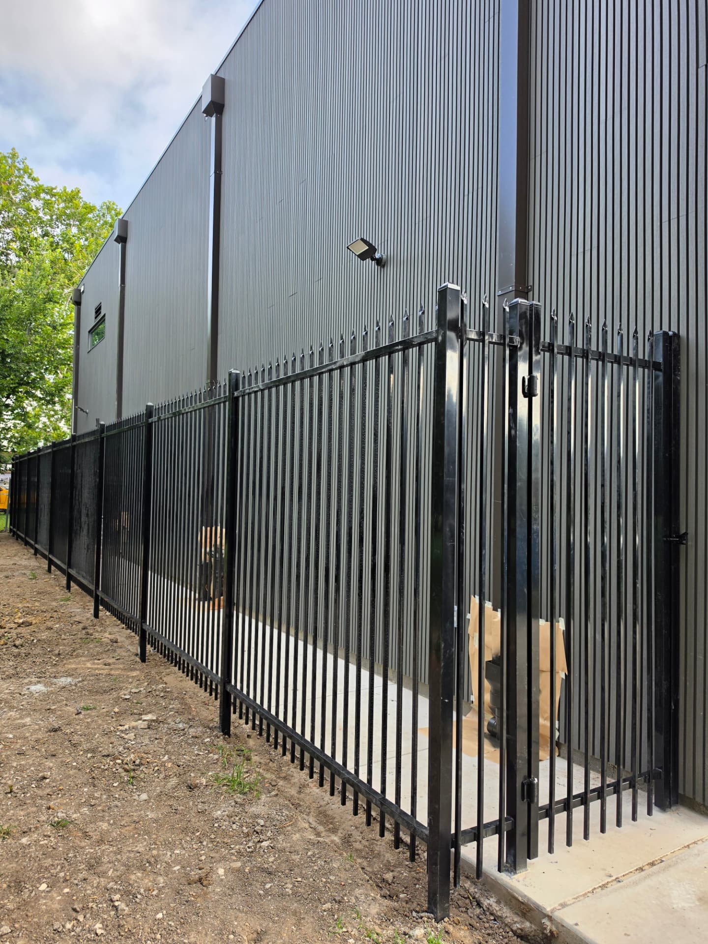Gloss black steel spear-top security picket fence with square posts and a swinging gate, running alongside a dark gray corrugated metal warehouse.