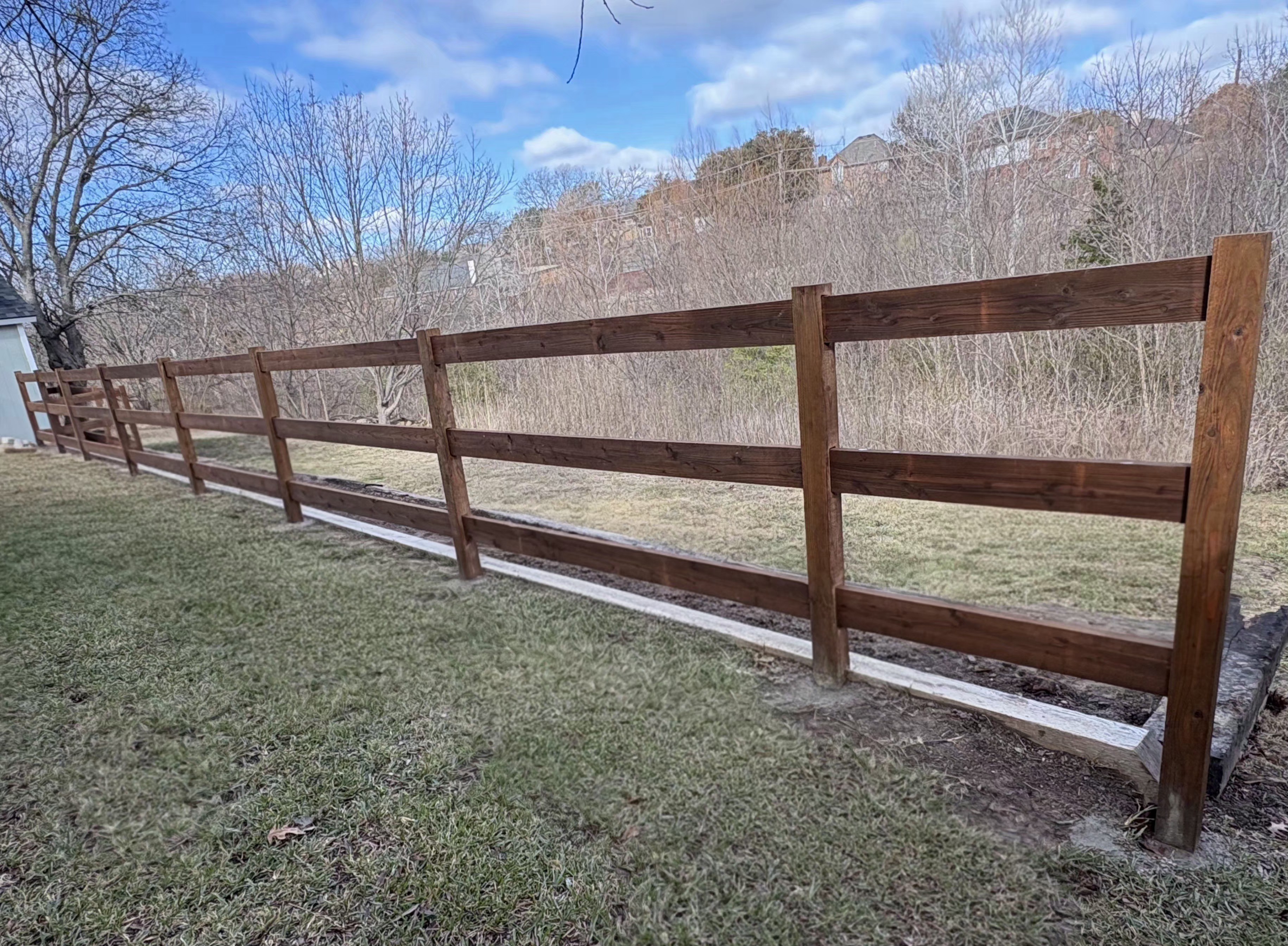 Three-rail board fence of wide flat boards stained deep warm brown on square wood posts with flat-cut tops and a light kick board along the ground, running along a yard that slopes down to leafless winter brush with rooftops on the ridge behind.