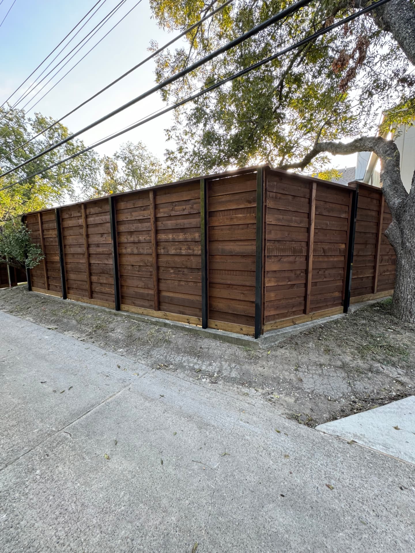 Horizontal slat cedar privacy fence in a dark walnut stain with exposed black steel posts and a lighter kick board, turning a corner along a concrete alleyway.