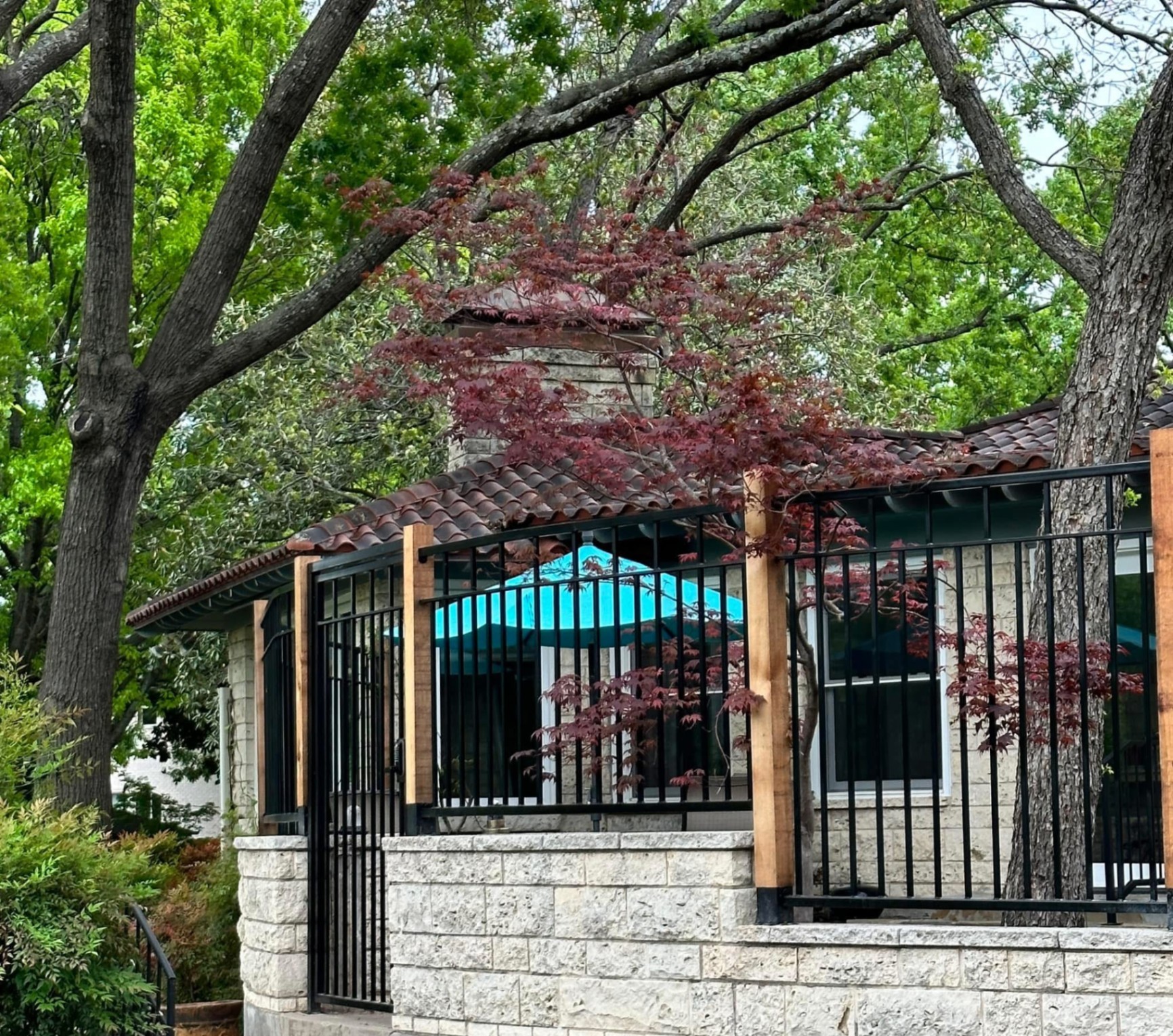 Matte black metal picket fence and matching walk gate mounted on a chiseled limestone retaining wall with natural wood posts, in a wooded backyard with a stone home and clay tile roof behind.