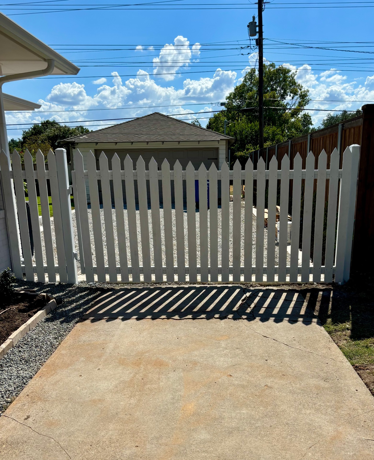 White picket-style driveway gate with a matching short picket fence, hung on metal hinges beside a stained wood privacy fence, opening onto a concrete driveway with a gravel lot and detached garage behind.