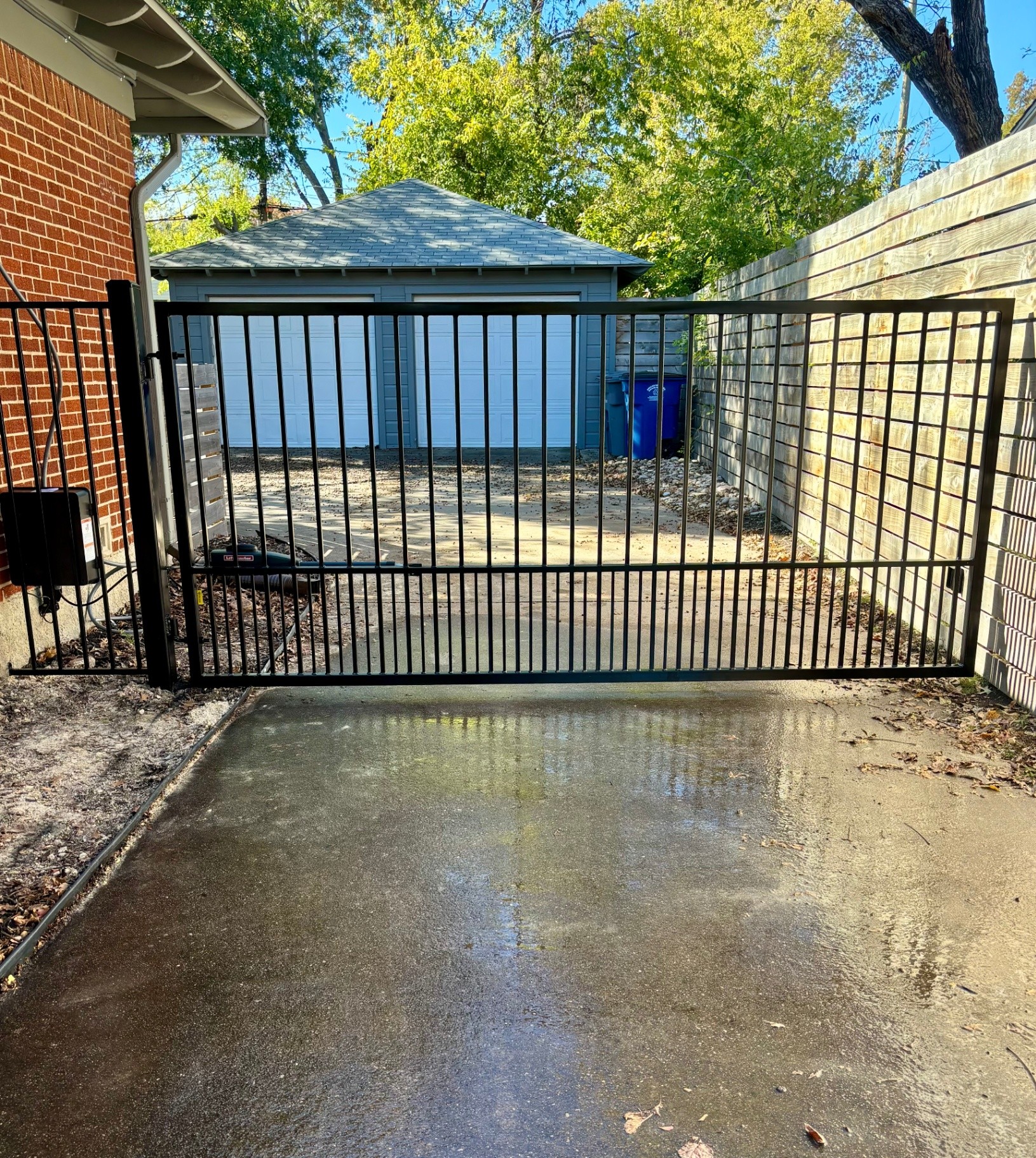 Black metal swing driveway gate with closely spaced puppy pickets along the bottom, a LiftMaster linear actuator operator and barrel hinges, set between a red brick wall and a horizontal wood slat fence.