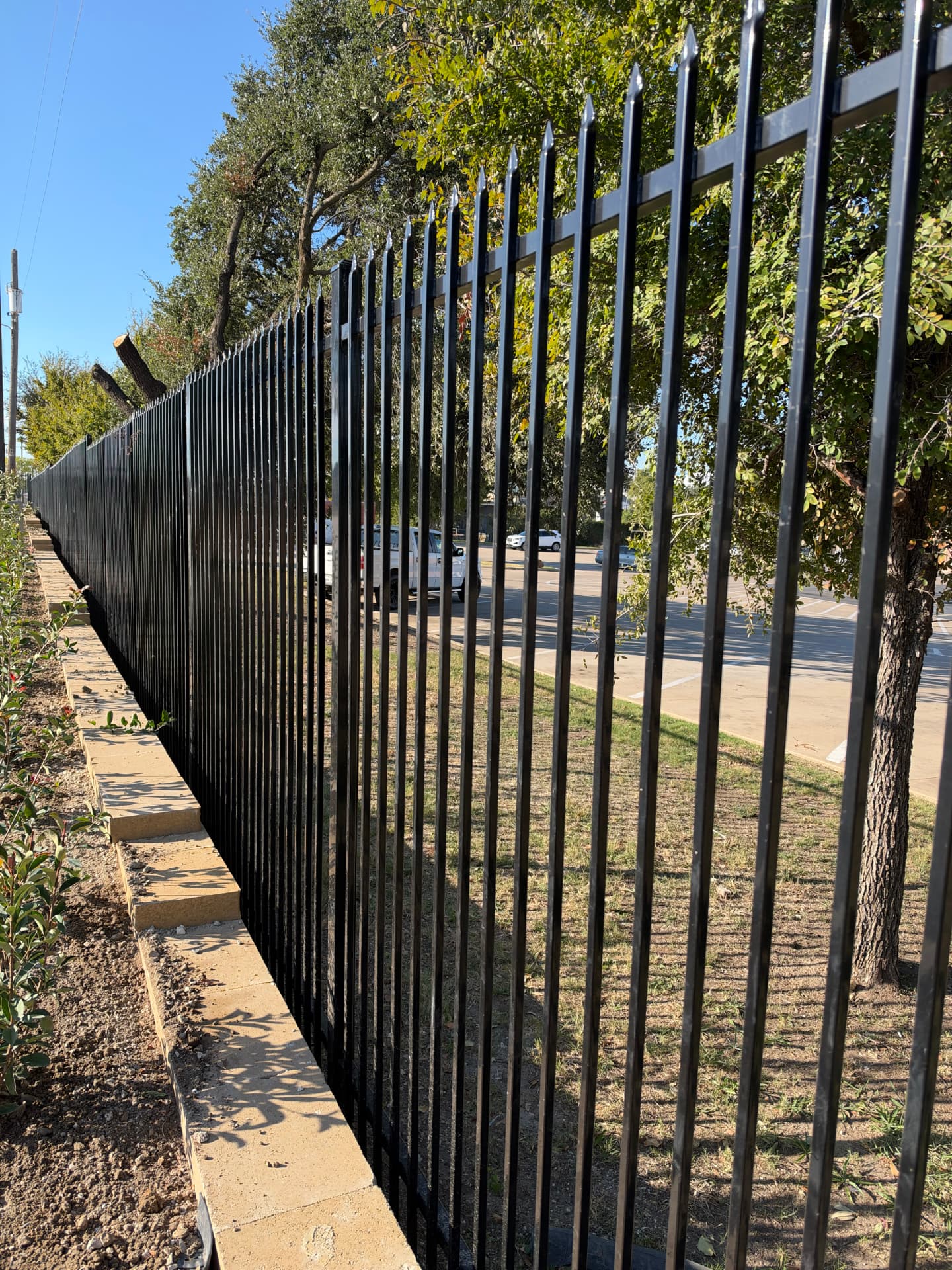 Black spear-top steel perimeter fence stepping down a concrete block retaining wall beside a commercial parking area.