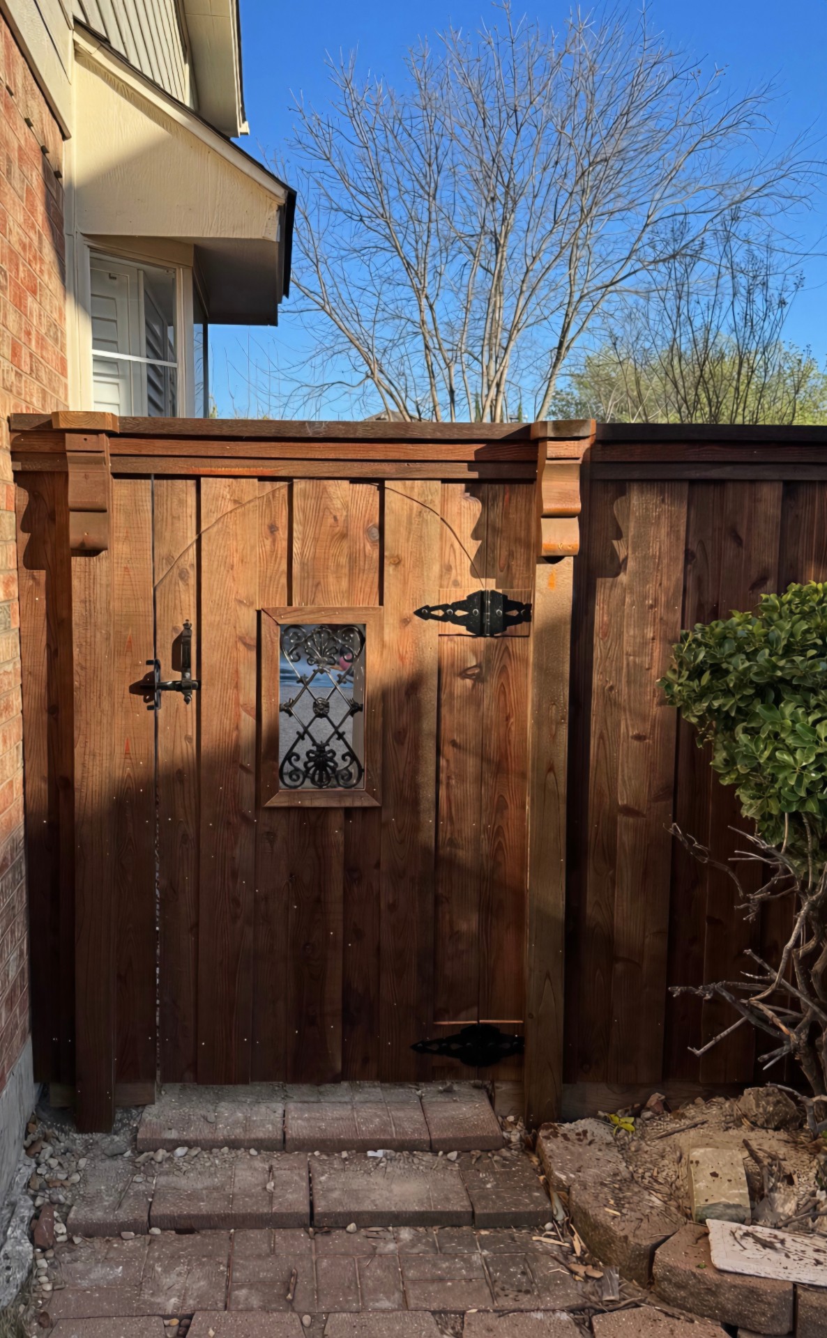 Vertical cedar plank privacy fence in a warm amber stain with a cap rail and carved wood corbels on the gate posts, framing a board gate with an arched routed relief, an ornate black iron scroll grille over a speakeasy opening, black strap hinges and a black latch.