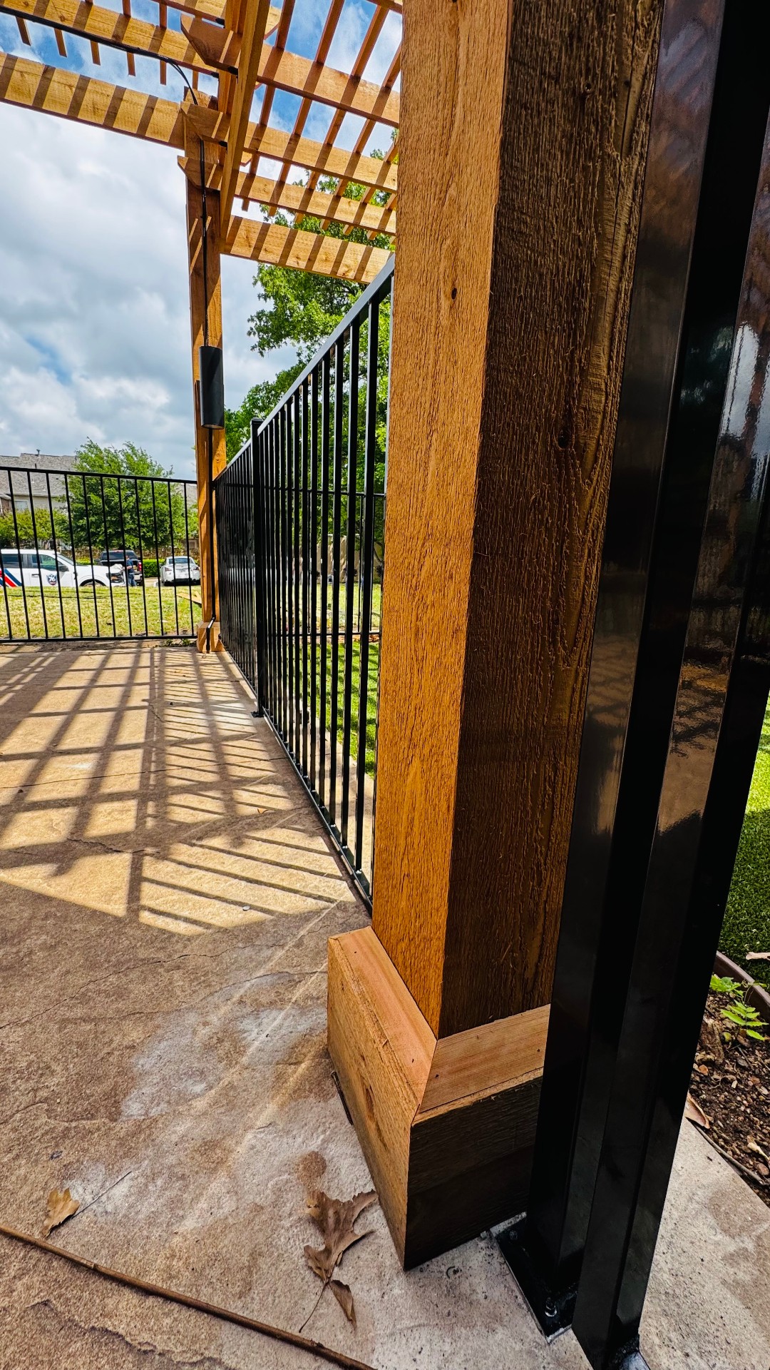 Glossy black metal picket fence panels with square posts mounted on base plates, flanking a stained timber pergola column over a stamped concrete patio with artificial turf beyond.