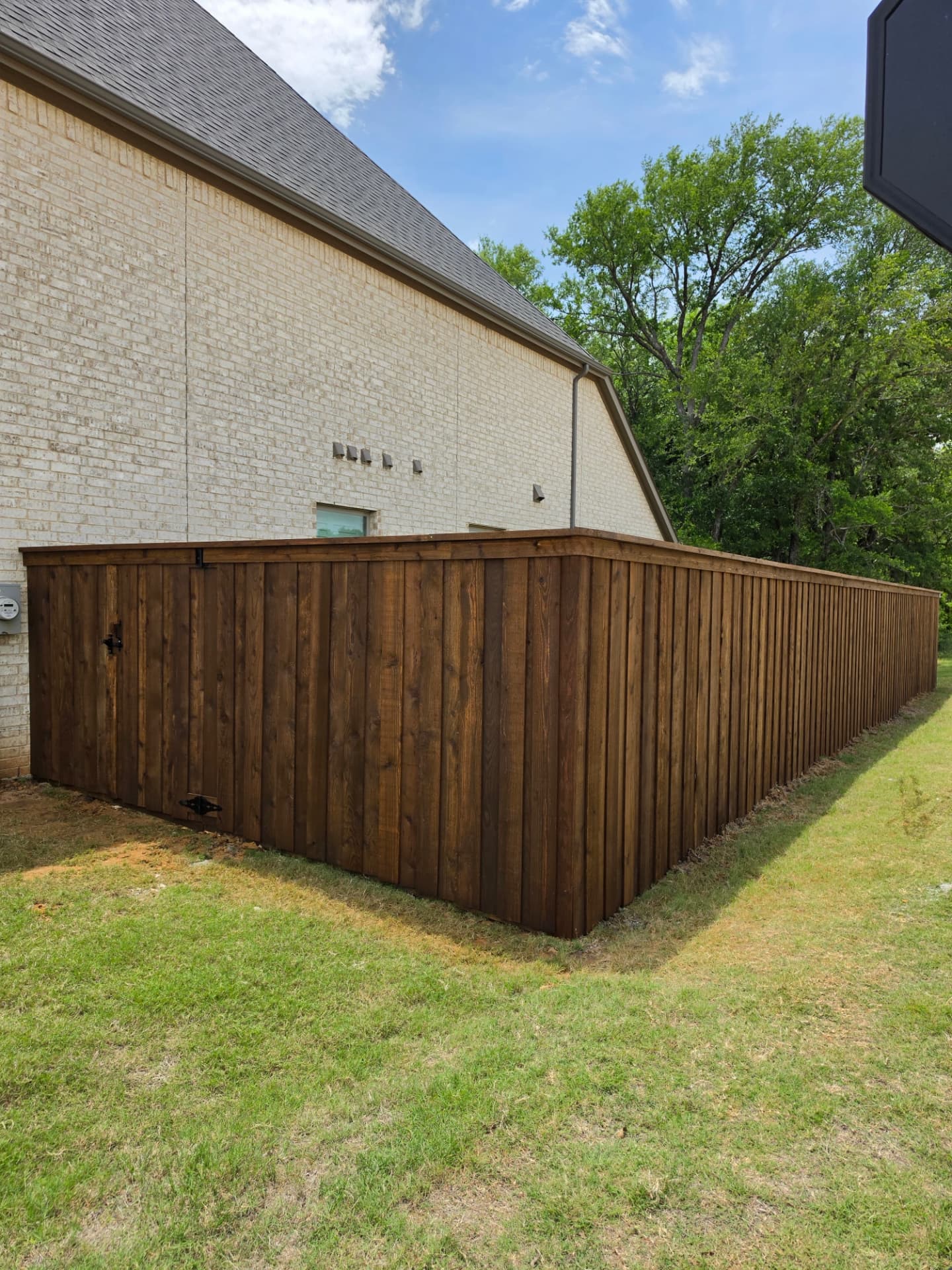 Board-on-board cedar privacy fence in a rich brown stain with a cap board and a matching gate hung on black strap hinges, beside a light brick home.
